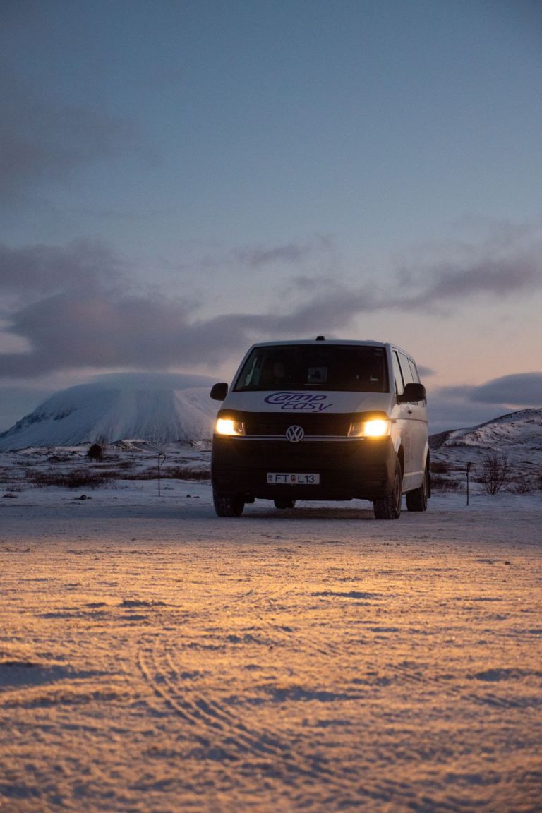Camper van with light on parked on the snowy parking with the snowy mountains in the background.