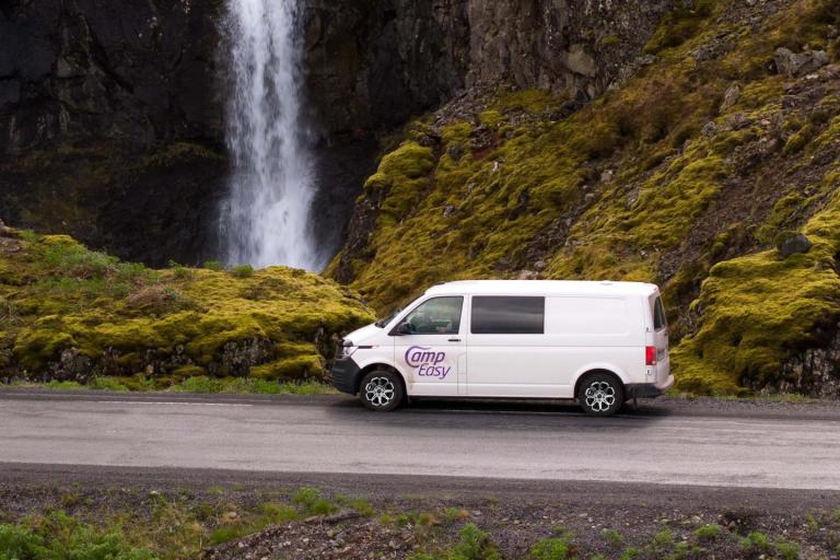 White campervan with "CampEasy" logo parked in Iceland near a stunning waterfall and moss-covered cliffs.