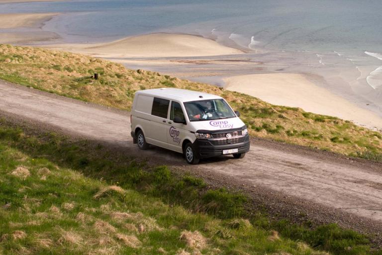 White camper van with 'Camp Easy' logo driving on a dirt road near a sandy beach in Iceland.