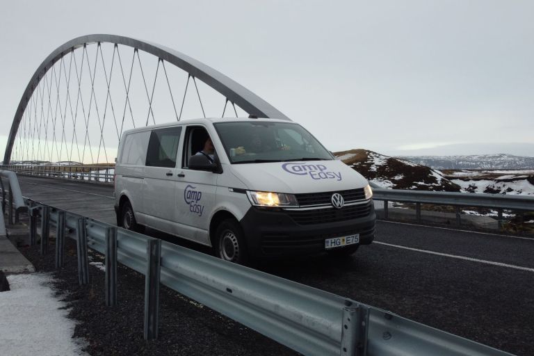 White camper van with "CampEasy" logo on bridge with arched structure in Iceland, surrounded by snowy hills.
