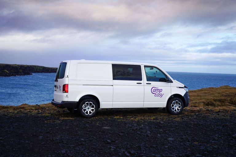 White 'Camp Easy' camper van parked on a rugged black gravel path overlooking a stunning ocean coastline with gentle waves, framed by a dramatic cliffside and distant mountains under a vast, colorful sky with soft clouds at sunrise or sunset in Iceland.