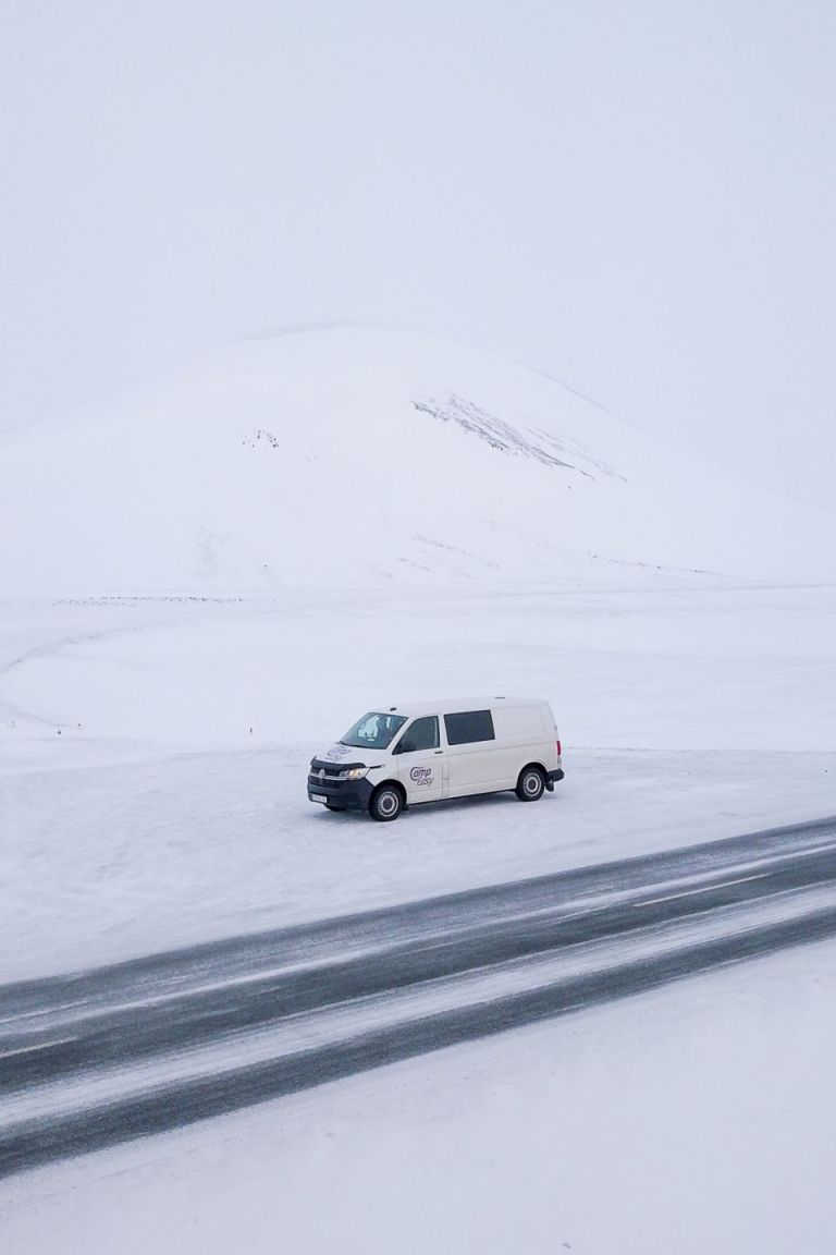 A white camper van parked next to the road, surrounded by snow, and a barely visible mountain covered by snow.