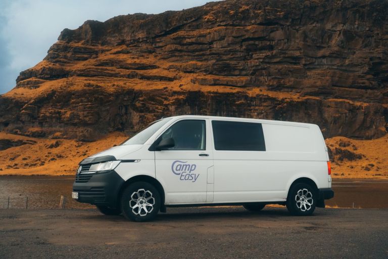 White 'Camp Easy' campervan parked near a lake with rocky cliffs in the background.
