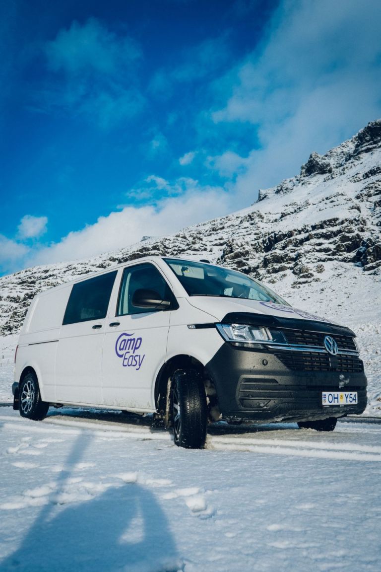 White 'Camp Easy' campervan parked on a snowy road with mountains under a blue sky in Iceland.