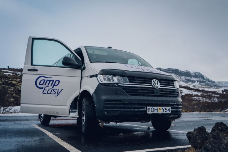 White 'Camp Easy' campervan with an open door parked in a snowy lot with mountains in the background.