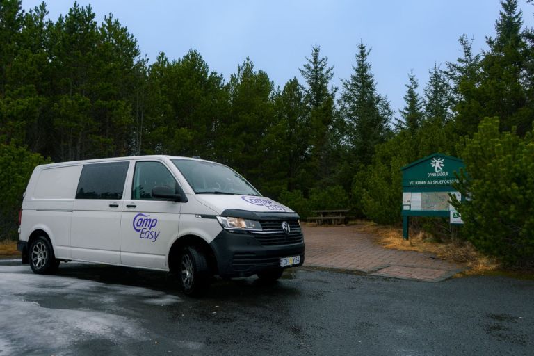 A campervan is parked on the road next to the wooden bench, sign, and dark green trees.