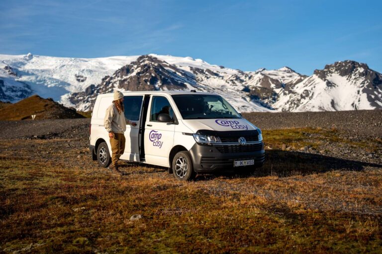 A man is closing the side doors of the campervan parked on the grass with the mountains in the background.