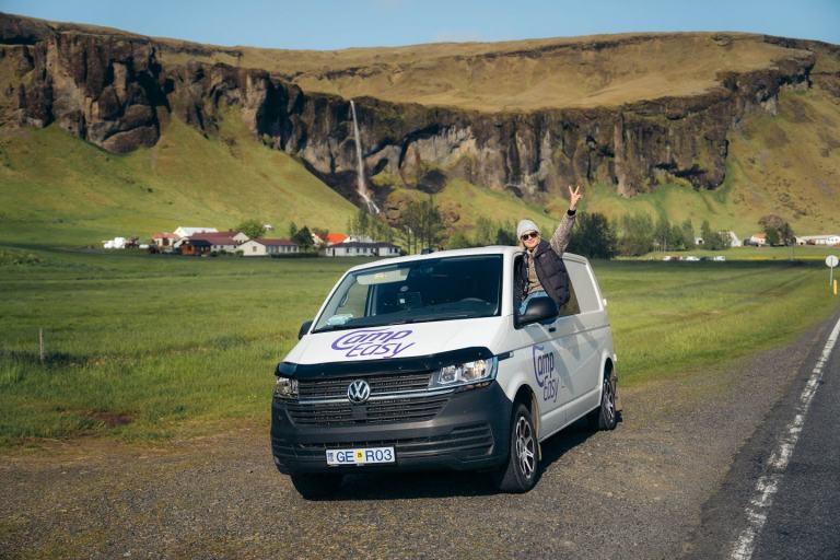 A woman is leaning out of the campervan window. Campervan is parked on the roadside next to the green grass field, building and waterfall.