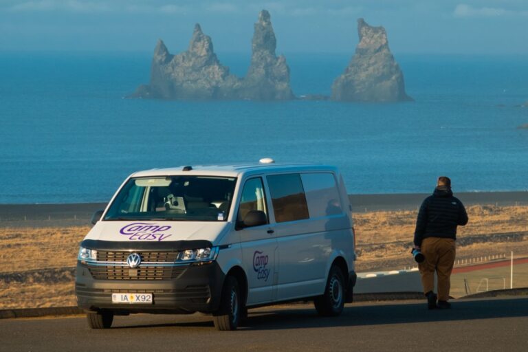 A man is walking next to the campervan parked in the parking lot. In the background there are famous Icelandic rock formations.