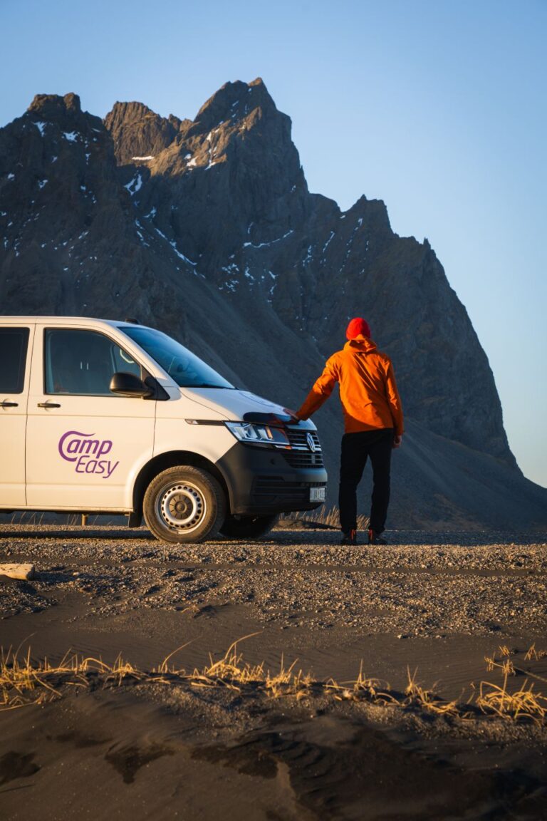 A man with a red cap and an orange jacket is leaning on the front of the parked campervan with the huge mountain in the background.