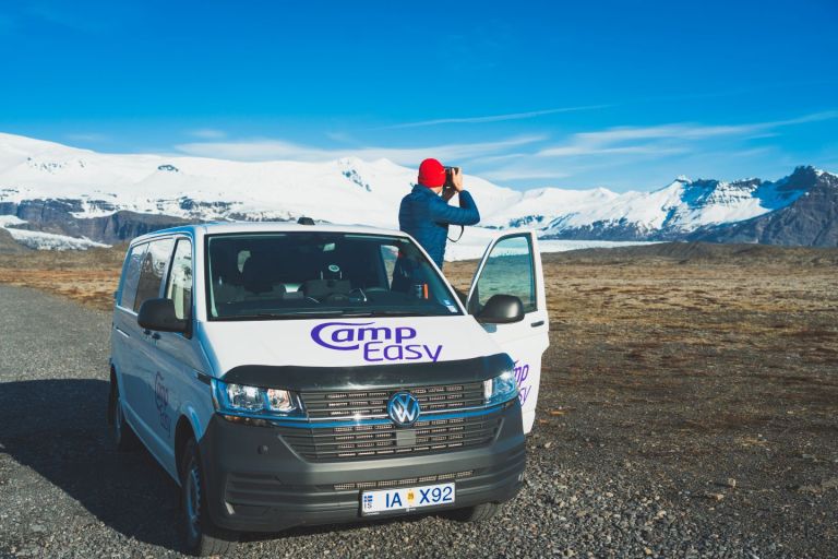 A man with a rad cap and a blue jacket is standing next to the camper van and taking a picture of the snowy mountains.