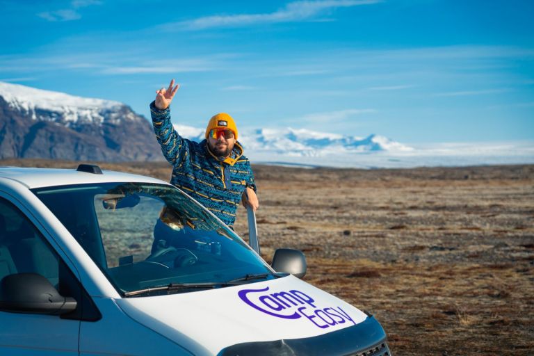 Man with yellow cap standing on white campervan with "CampEasy" logo in Iceland, with snowy mountains and clear sky.