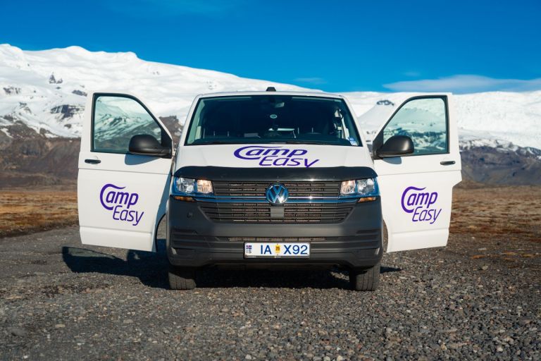 A white campervan with opened front doors parked on the gravel road with the snowy mountains in the background.
