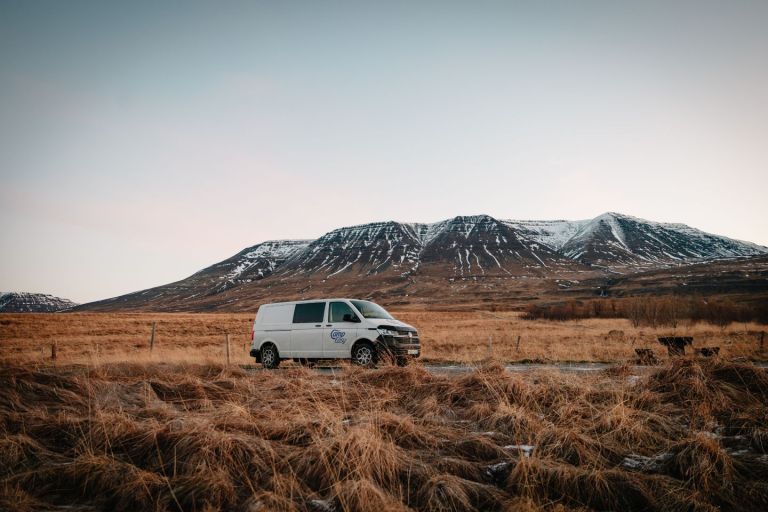 Campervan parked on the road next to the wooden bench and brown grass fields with the snowy mountains in the background.