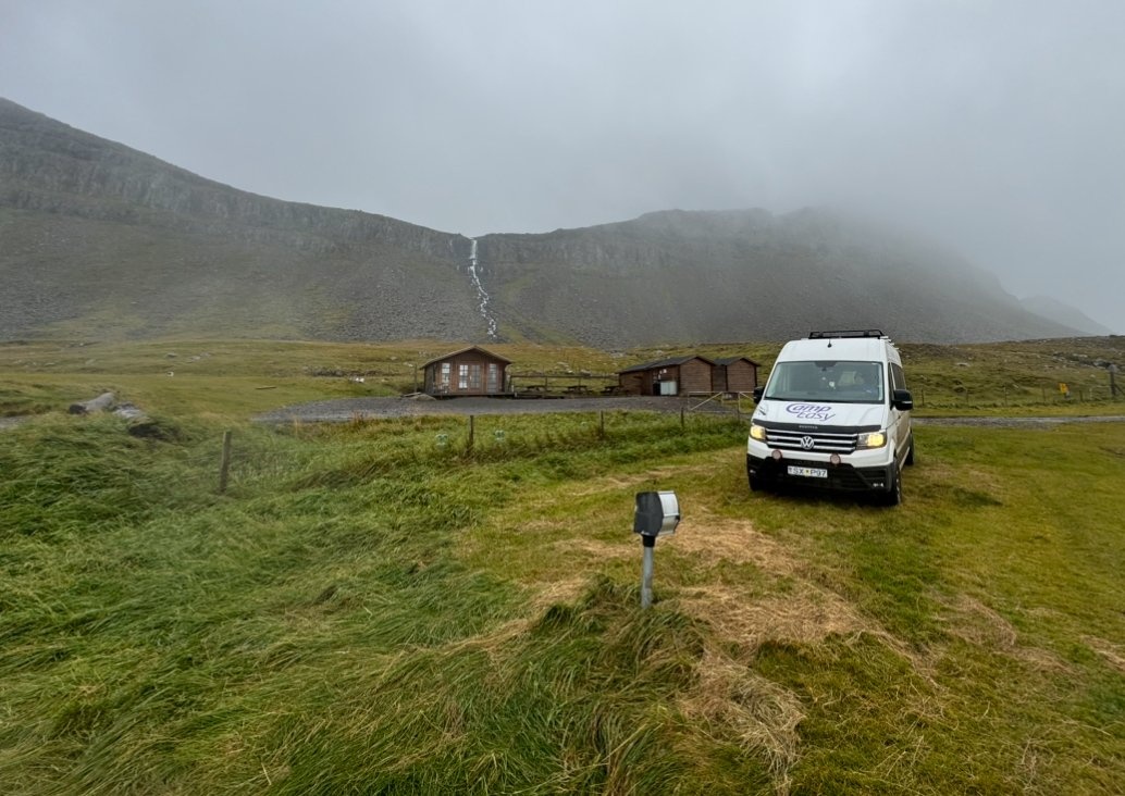 CampEasy campervan parked near small cabins with a dramatic waterfall cascading down a vast mountain in the background, under misty skies, capturing the wild and natural beauty of Iceland.