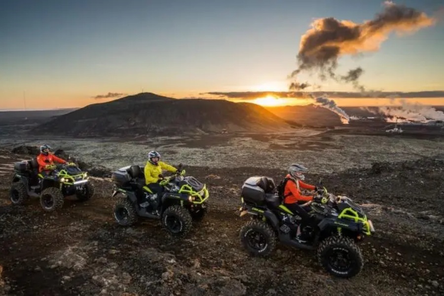 Three people riding ATVs across a rugged volcanic landscape in Iceland during sunset, with steam rising from distant geothermal vents and a mountainous horizon in the background.