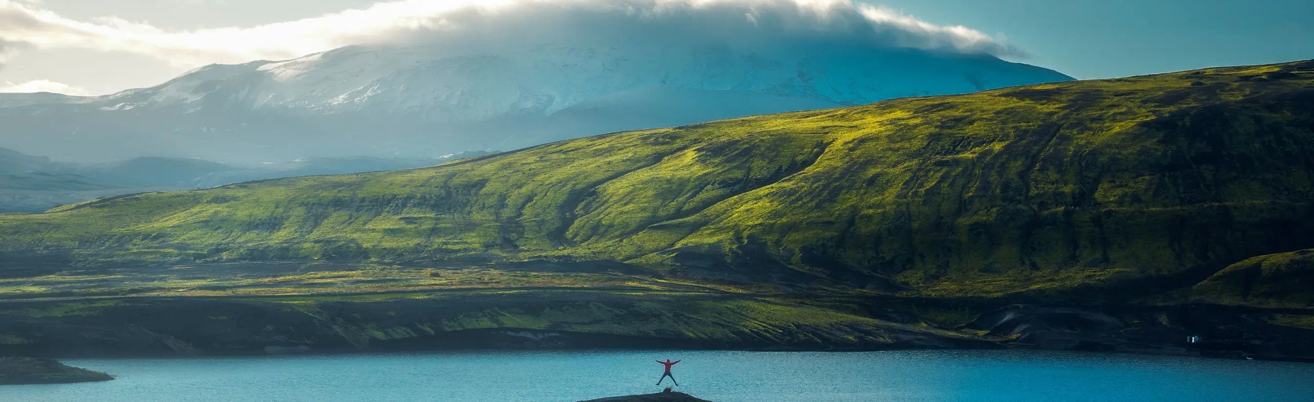 Person standing with arms open by a lake, surrounded by green hills and misty snow-capped mountains in the background.