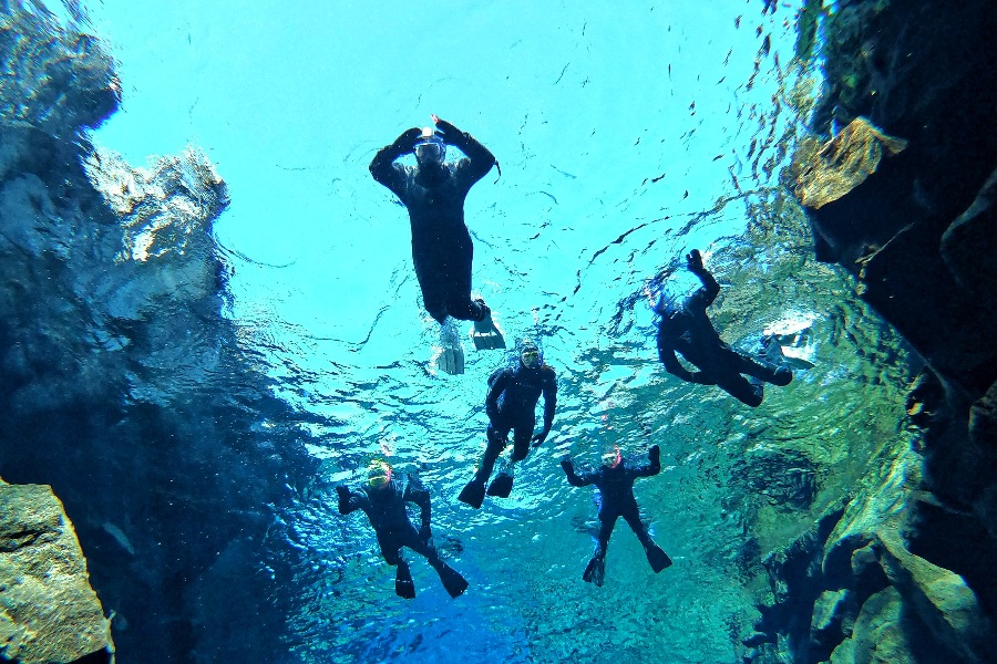 A group of five snorkelers in wetsuits swimming in the clear blue waters of Iceland's Silfra fissure, surrounded by rocky underwater cliffs with sunlight illuminating the scene from above.