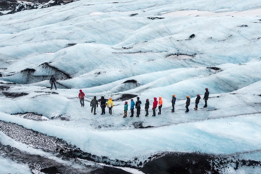 A group of hikers in colorful jackets and helmets trekking across the icy landscape of the Sólheimajökull glacier in Iceland, with the rugged, crevassed surface of the glacier stretching out around them.