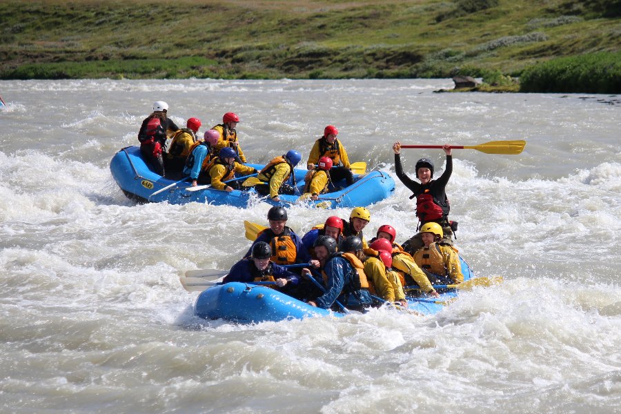 Two groups of people in blue rafts paddling through whitewater rapids on a river in Iceland, wearing helmets and life jackets, with one person in the front raft triumphantly raising a paddle in the air.