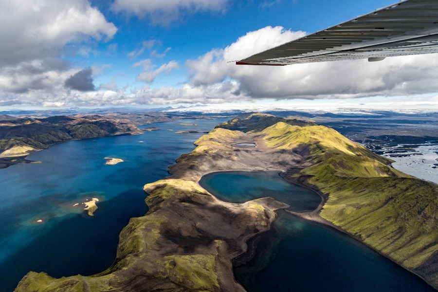 An aerial view of Iceland's rugged landscape with vibrant blue lakes surrounded by green and brown mountains, as seen from the window of a small airplane, with part of the wing visible in the upper right corner of the image.