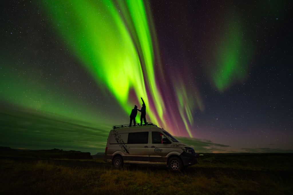 Two people standing on top of a CampEasy camper van under the Northern Lights, with vibrant green and purple aurora illuminating the night sky in Iceland.