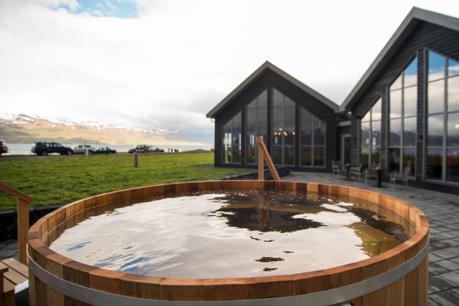 A wooden outdoor hot tub filled with water, located outside a modern building with large windows, set against a backdrop of grassy fields and distant mountains in Iceland.