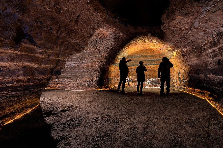 Three people silhouetted against the warm lighting inside a large underground cave, examining the textured rock walls of the Caves of Hella in Iceland.