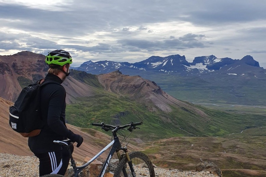 A cyclist wearing a green helmet and black gear stands beside their mountain bike, gazing out over a vast, rugged Icelandic landscape with towering mountains and green valleys stretching into the distance under a cloudy sky.