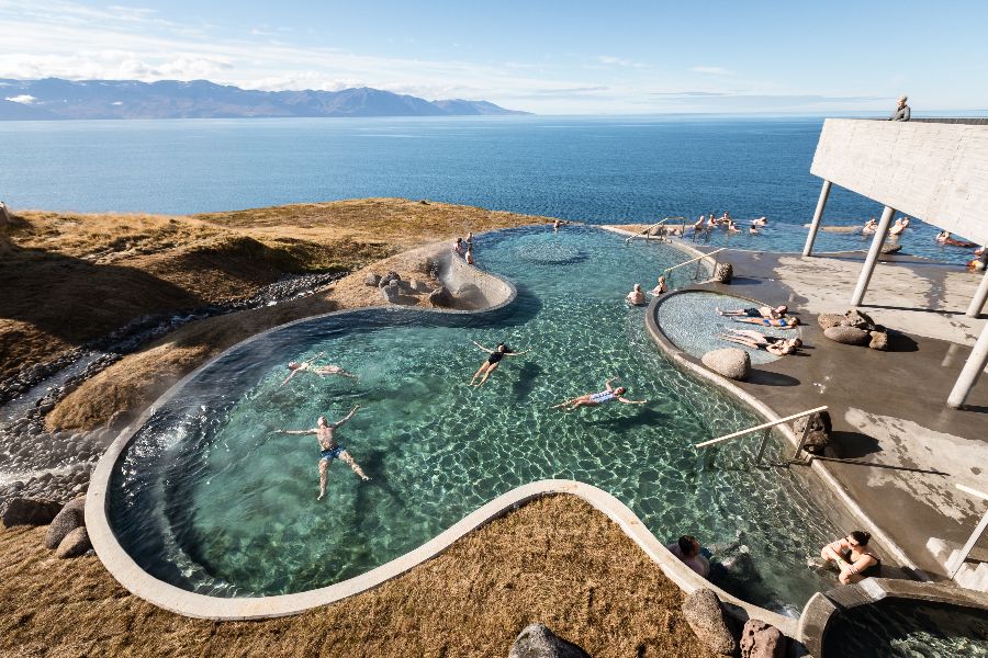 Visitors relaxing in the naturally heated outdoor infinity pools of GeoSea, overlooking the vast blue ocean and distant mountains on a clear day in Iceland, with some people floating in the warm geothermal waters and others lounging on the poolside deck.