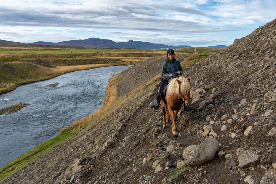 A person riding an Icelandic horse along a rocky hillside trail next to a river, with expansive fields and distant mountains visible under a partly cloudy sky in the background.