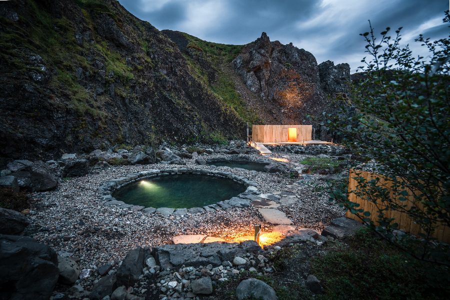 A peaceful natural hot spring pool at the Húsafell Canyon Baths in Iceland, surrounded by rocky cliffs and illuminated by soft lighting, with a small wooden building in the background under a cloudy evening sky.