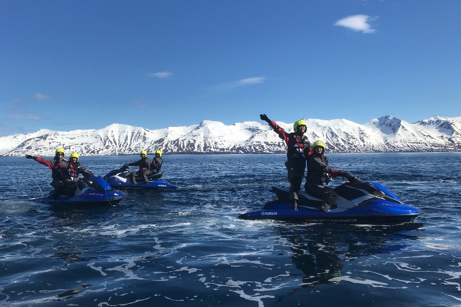 Group of people on blue Yamaha jet skis enjoying a tour on a clear day with snowy mountains in the background, wearing safety gear and helmets, with one person on the front jet ski raising their arms in excitement.