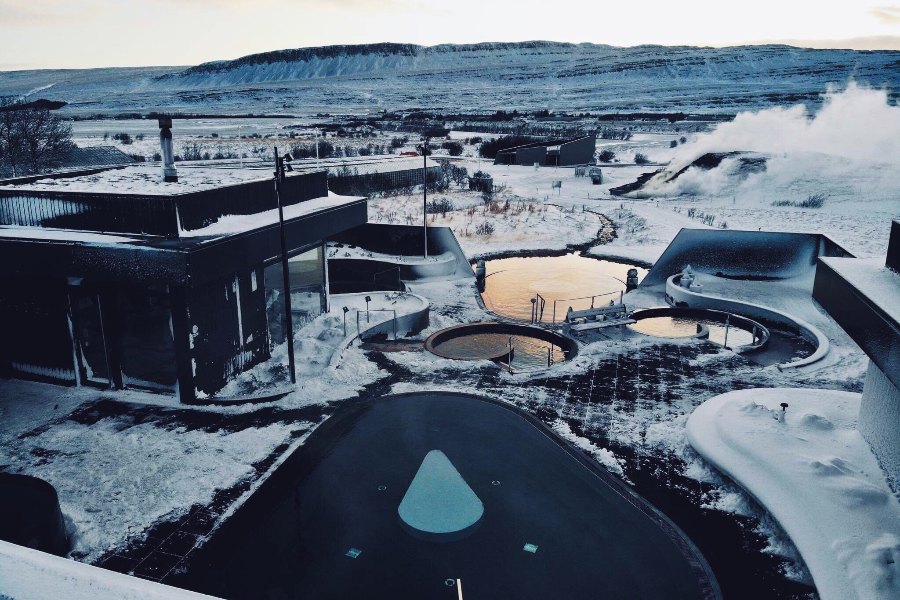 A view of the Krauma geothermal baths in Iceland during winter, with outdoor hot pools surrounded by snow-covered walkways and buildings, steam rising from the hot springs in the distance.