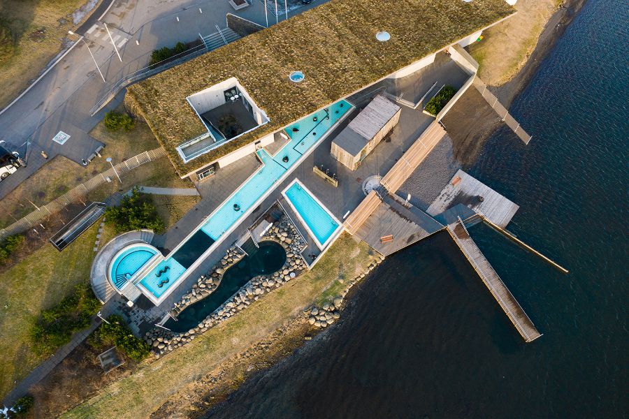 An aerial view of the Laugarvatn Fontana geothermal baths, featuring outdoor pools and hot tubs, a wooden boardwalk, and a building with a grass-covered roof, located next to the edge of a lake in Iceland.