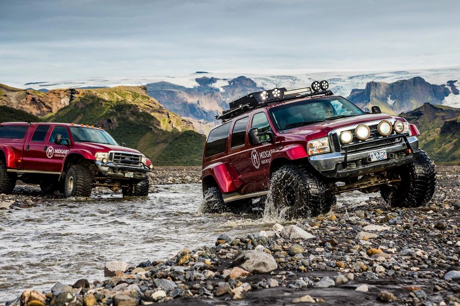 Two large red super jeeps from Midgard Adventure crossing a shallow river in Iceland’s rugged terrain, with rocky mountains and glaciers visible in the background.