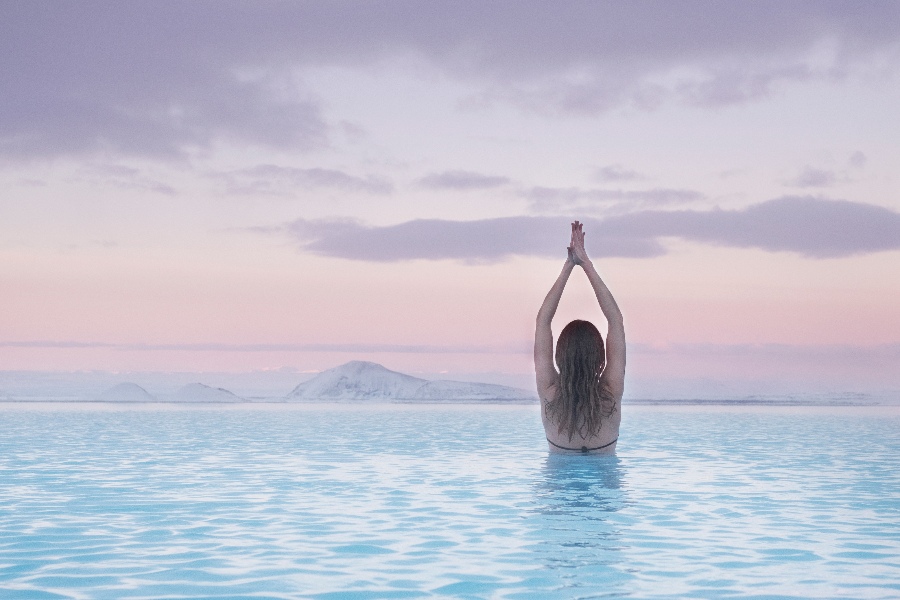 A woman standing in the serene blue waters of the Mývatn Nature Baths in Iceland, facing the horizon with her arms raised in a yoga pose, surrounded by snow-capped mountains and a pastel-colored sky at sunrise or sunset.