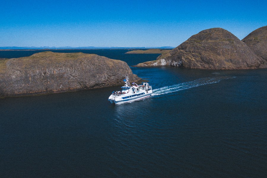 A white and blue tour boat cruising through calm waters between two rocky islands during the Viking Sushi Tour in Iceland, with clear blue skies overhead.