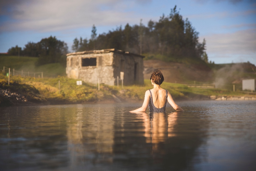 A woman wading in the warm waters of the Secret Lagoon in Iceland, facing an old stone building and lush green landscape, with steam rising from the geothermal pool under a soft, cloudy sky.