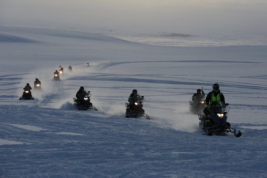 A group of snowmobilers riding in formation across a vast snowy landscape, with their headlights cutting through the foggy, overcast atmosphere as they leave tracks in the snow.