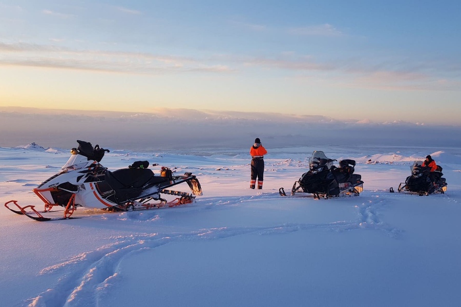 Several snowmobiles parked on a snowy landscape during a serene sunset. People wearing bright orange jackets stand nearby, gazing out at the vast horizon with soft clouds in the distance.