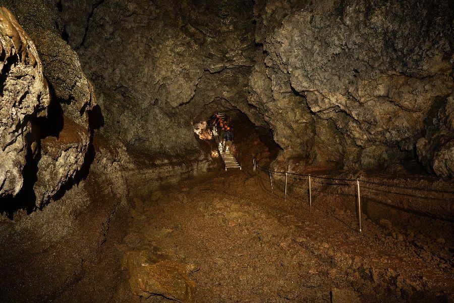 A group of explorers walking through a dark, rocky cave passage with uneven walls, guided by a rope and illuminated by a faint light, as they ascend a narrow staircase deeper into the cave.