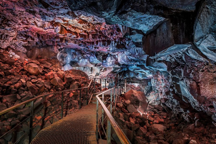 A vibrant, illuminated walkway inside a lava tunnel, with the rugged rock walls glowing in shades of red, purple, and blue, guiding visitors through the dramatic volcanic cave filled with large boulders and rock formations.