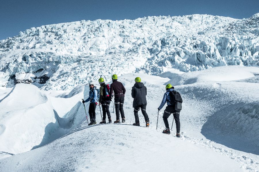 A group of hikers with helmets and trekking poles stand on a glacier, surrounded by vast snowfields and an enormous ice wall in the background. They are taking in the breathtaking view of the frozen landscape.