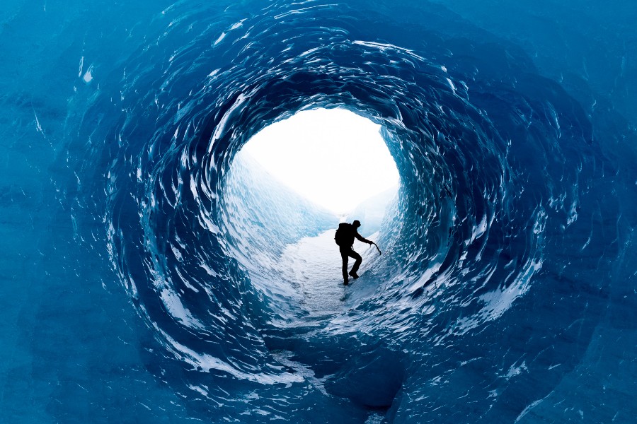 A lone explorer silhouetted inside a tunnel of vibrant blue ice in Vatnajökull, Iceland, walking toward the bright light at the end of the cave, surrounded by smooth, frozen walls.