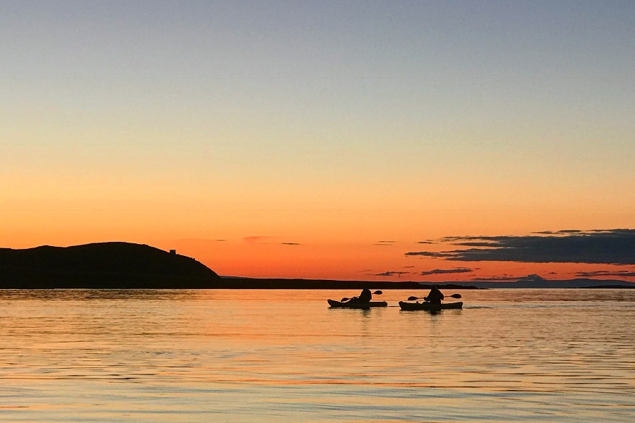 Two kayakers paddling across calm waters during a vibrant sunset, with the sky transitioning from orange to blue and a silhouette of a distant hill on the horizon.