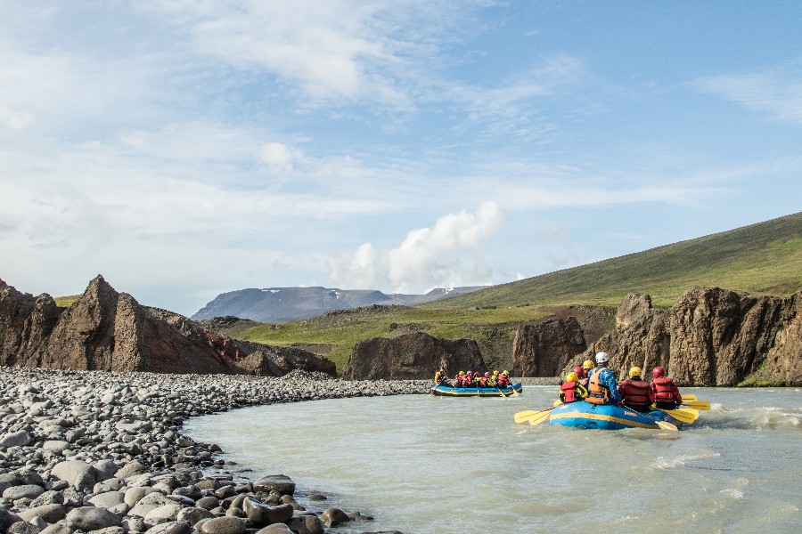 Two groups of people in yellow and blue rafts navigating a glacial river through a rocky landscape in Iceland, surrounded by green hills and distant mountains under a partly cloudy sky.