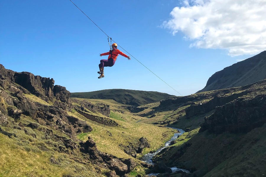 A person ziplining across a deep green valley in Iceland, with rocky cliffs and a winding stream below, under a bright blue sky.