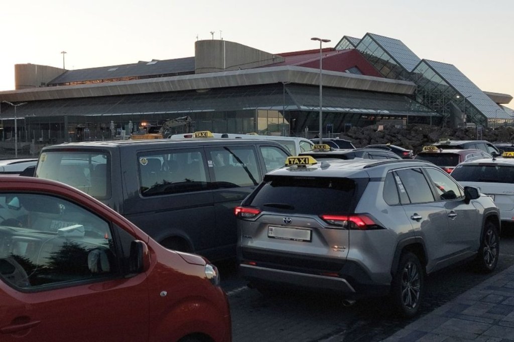 The image shows a row of taxis lined up outside Keflavík International Airport in Iceland. The modern airport building with glass and geometric architectural features serves as the backdrop. The scene appears to be during early morning or evening hours, as indicated by the low sunlight.