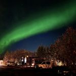 Stunning view of the aurora borealis streaking across the sky in shades of green above a campsite, with trees and a parked van beneath a star-filled sky.